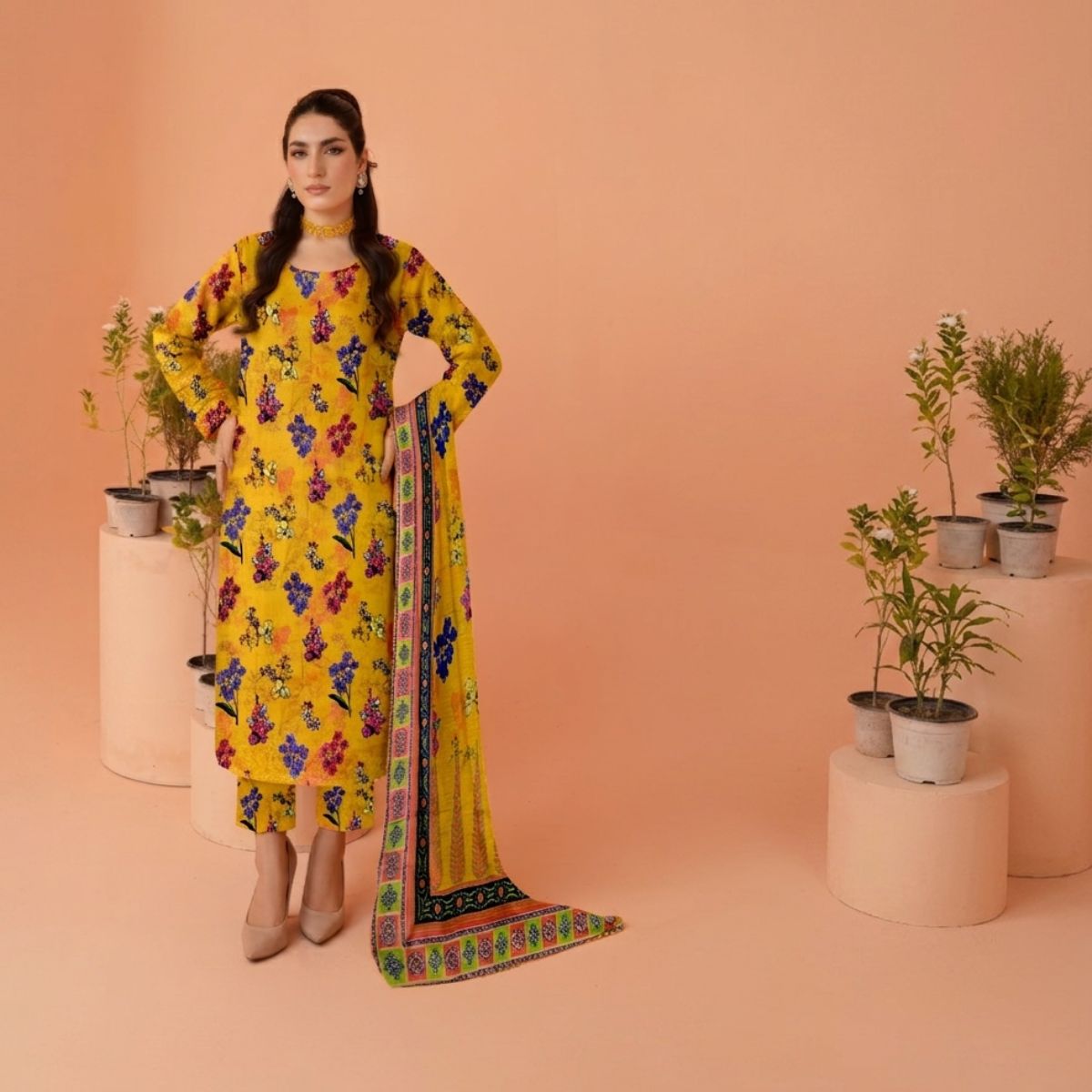 Woman in a yellow floral dress with a colorful scarf standing against a peach background with potted plants.